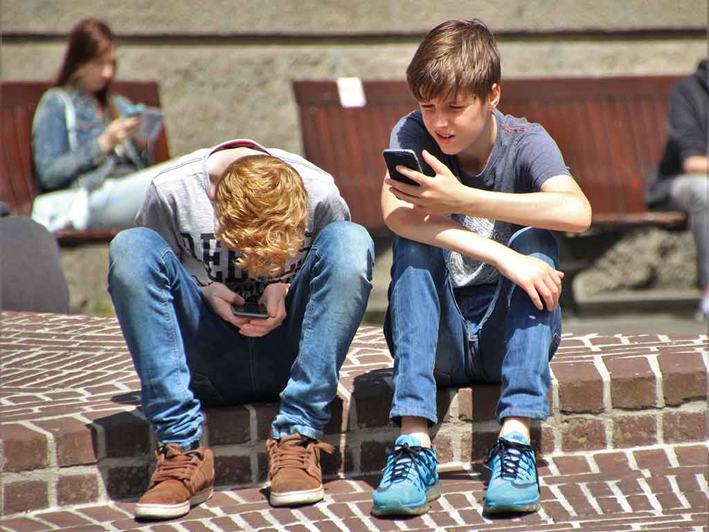 Das Foto zeigt zwei Jungs im Teenager-Alter, die draußen über Ihre Handys gebeugt sitzen und auf den Smartphones spielen, als Vorschau für den medienpädagogischen Eltern-Workshop.