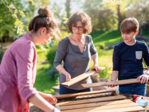 Das Foto zeigt eine Naturpädagogin, die mit zwei Teenagern im Freien mit Holz arbeitet, es dient als Vorschau für den Permakultur-Workshop.